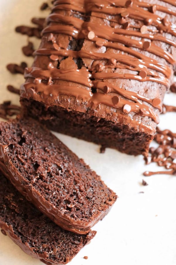 A sliced loaf of chocolate bread, topped with a drizzle of chocolate frosting, is displayed on a light-colored surface.
