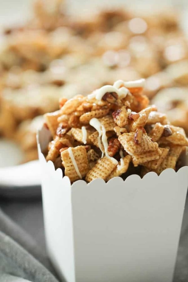 A close-up of a white container filled with caramel-coated snack mix, featuring cereal pieces and pretzels, drizzled with a white icing.