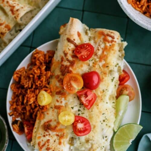 A plate with an enchilada topped with melted cheese and sliced cherry tomatoes, accompanied by a side of Mexican rice and a wedge of lime.