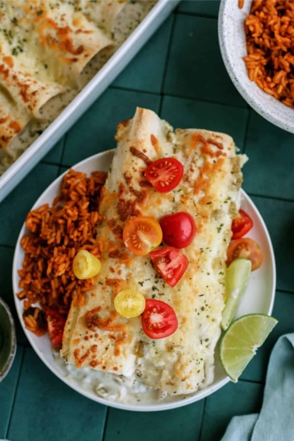 A plate with an enchilada topped with melted cheese and sliced cherry tomatoes, accompanied by a side of Mexican rice and a wedge of lime.