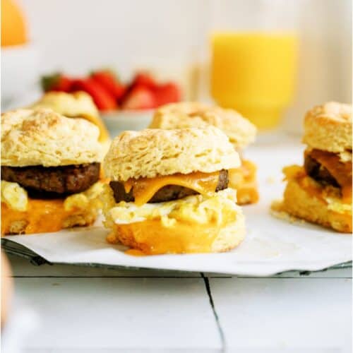 Breakfast sandwiches with sausage, egg, and cheese on biscuits, placed on parchment paper. In the background are a glass of orange juice and fresh strawberries.