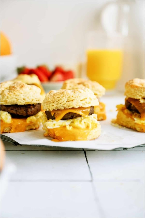 Breakfast sandwiches with sausage, egg, and cheese on biscuits, placed on parchment paper. In the background are a glass of orange juice and fresh strawberries.