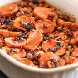 A white baking dish filled with sliced sweet potatoes, cranberries, and chopped nuts, baked and lightly glazed.