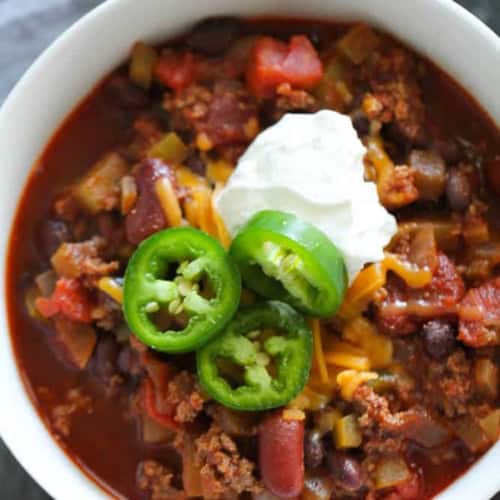 A bowl of chili topped with shredded cheese, sliced jalapeños, and a dollop of sour cream.