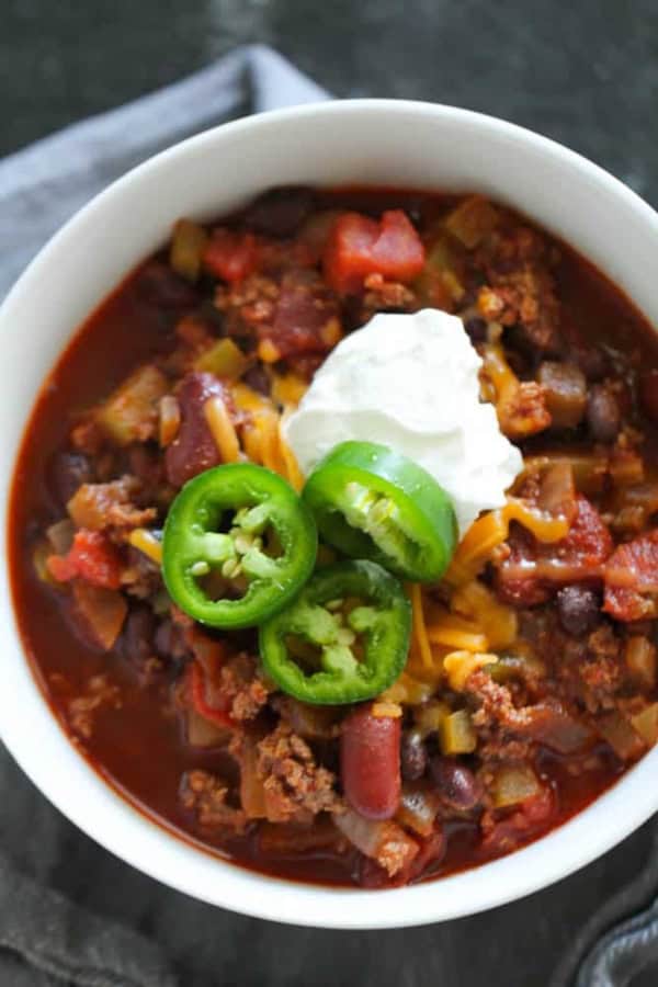 A bowl of chili topped with shredded cheese, sliced jalapeños, and a dollop of sour cream.