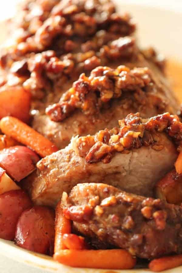 Sliced pork roast topped with a chopped nut glaze, served with cooked baby carrots and red potatoes.