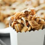 A close-up of a white container filled with caramel-coated snack mix, featuring cereal pieces and pretzels, drizzled with a white icing.