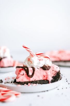 A slice of pink peppermint pie with a chocolate crust, topped with whipped cream, chocolate drizzle, and a peppermint stick, on a white plate.