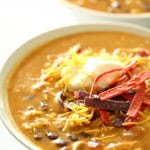 A bowl of creamy soup topped with shredded cheese, sour cream, and crispy strips, with another similar bowl in the background.