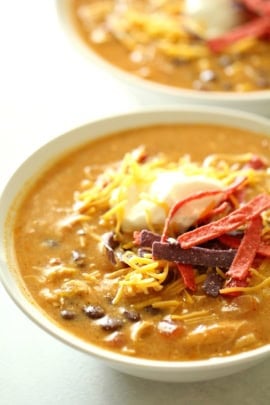 A bowl of creamy soup topped with shredded cheese, sour cream, and crispy strips, with another similar bowl in the background.