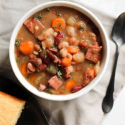 A bowl of bean and ham soup with carrots and herbs, next to a plate of cornbread and a black spoon on a beige napkin.