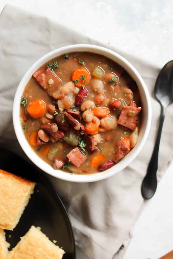 A bowl of bean and ham soup with carrots and herbs, next to a plate of cornbread and a black spoon on a beige napkin.