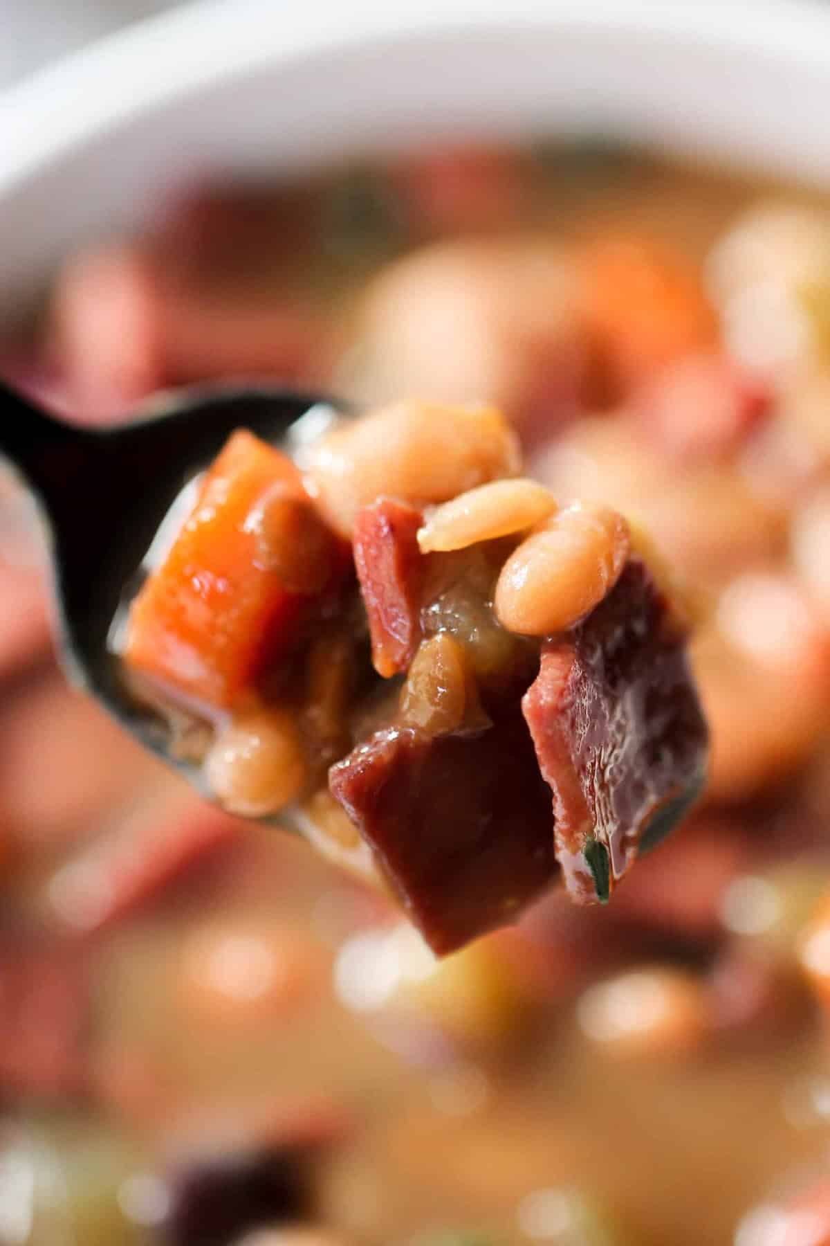 Close-up of a spoonful of soup containing beans, diced ham, carrots, and other vegetables in a broth.