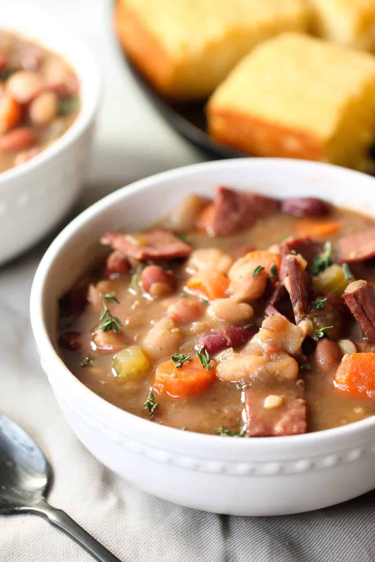 A bowl of hearty bean soup with diced vegetables and chunks of meat, garnished with herbs, served next to slices of cornbread.