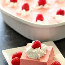 A square of pink gelatin dessert topped with whipped cream and a raspberry, served on a plate with extra raspberries, with the remaining dessert in a dish in the background.
