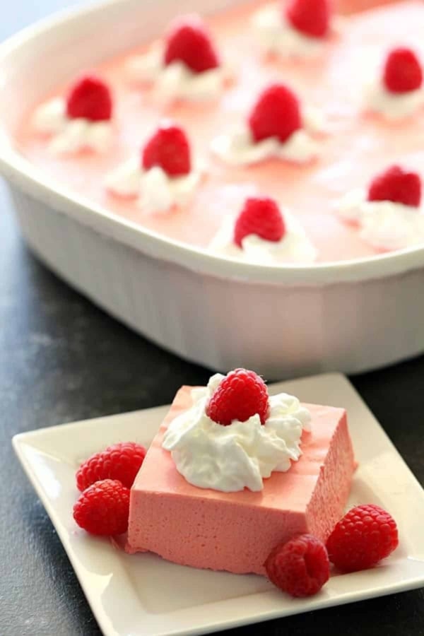 A square of pink gelatin dessert topped with whipped cream and a raspberry, served on a plate with extra raspberries, with the remaining dessert in a dish in the background.