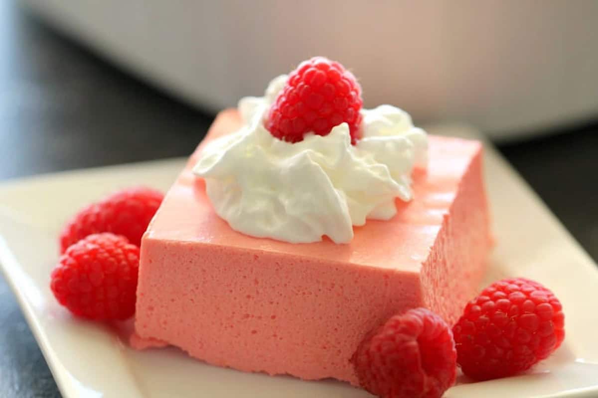 A square slice of pink mousse topped with whipped cream and a raspberry, garnished with additional raspberries on a white plate.