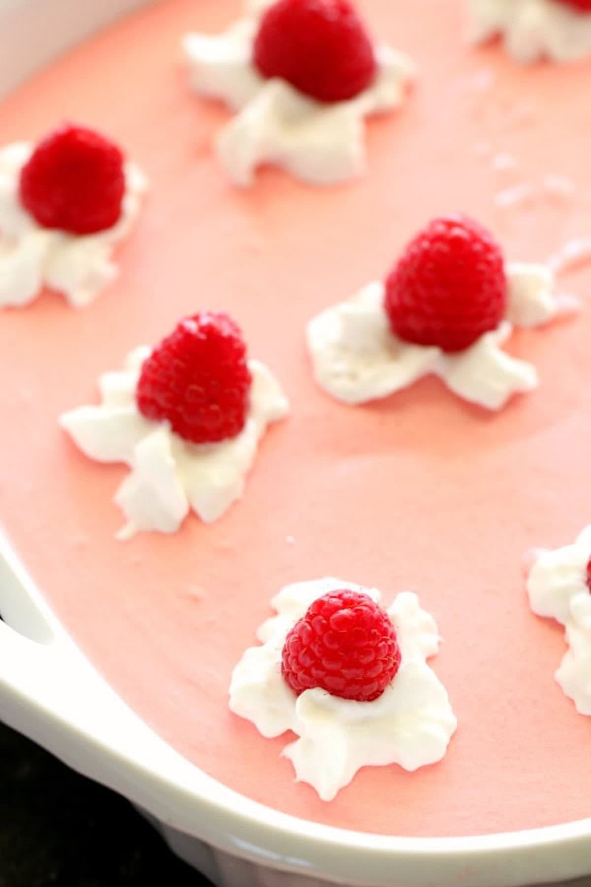A pink creamy dessert topped with dollops of whipped cream and fresh raspberries in a white dish.
