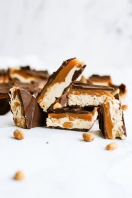 Several chocolate-covered caramel and nougat bars with peanuts, some cut in half, are displayed on a white surface.