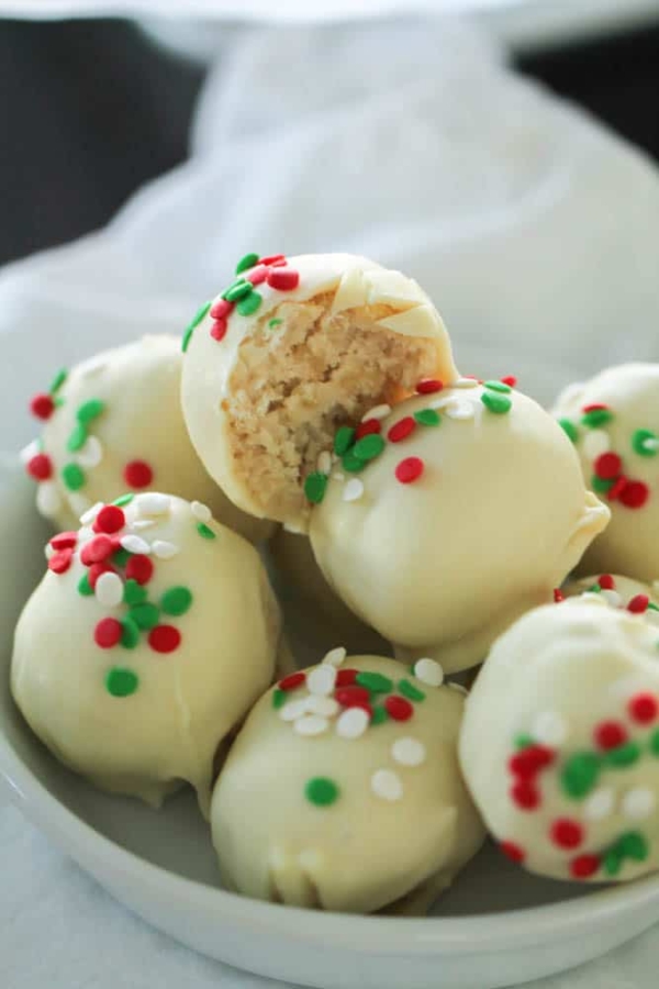 A bowl of white chocolate truffles decorated with red, white, and green sprinkles. One truffle has a bite taken out, revealing its crumbly interior.