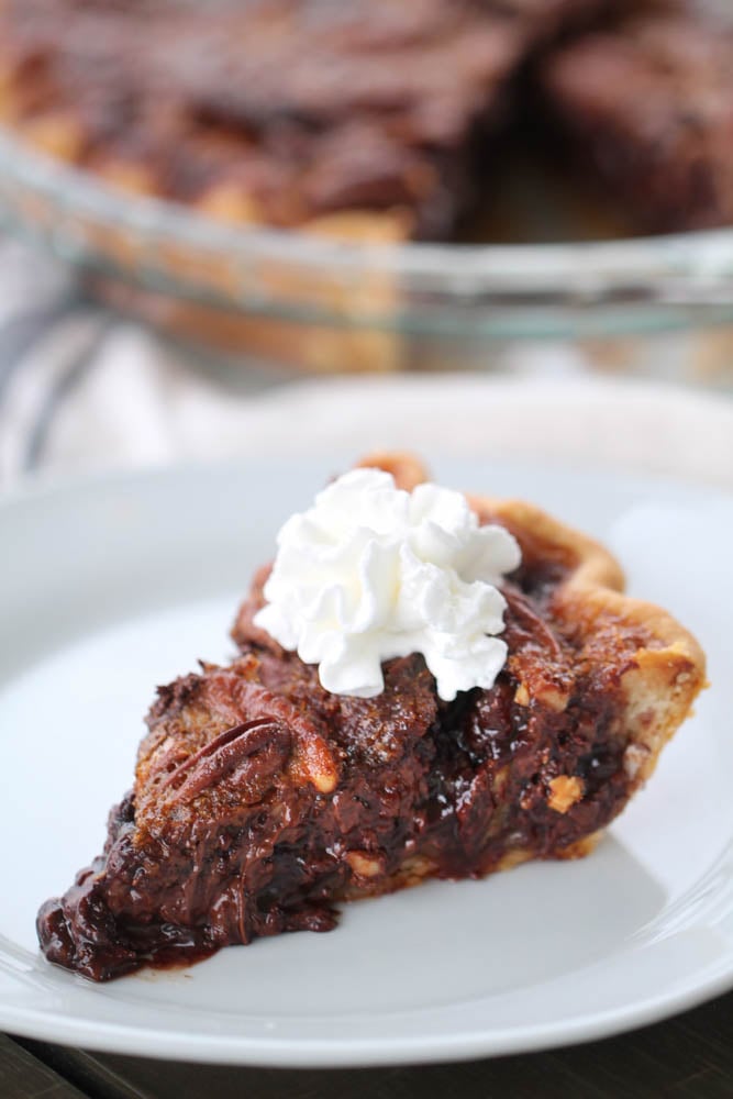 A slice of Chocolate Pecan Pie on a white plate topped with whip cream