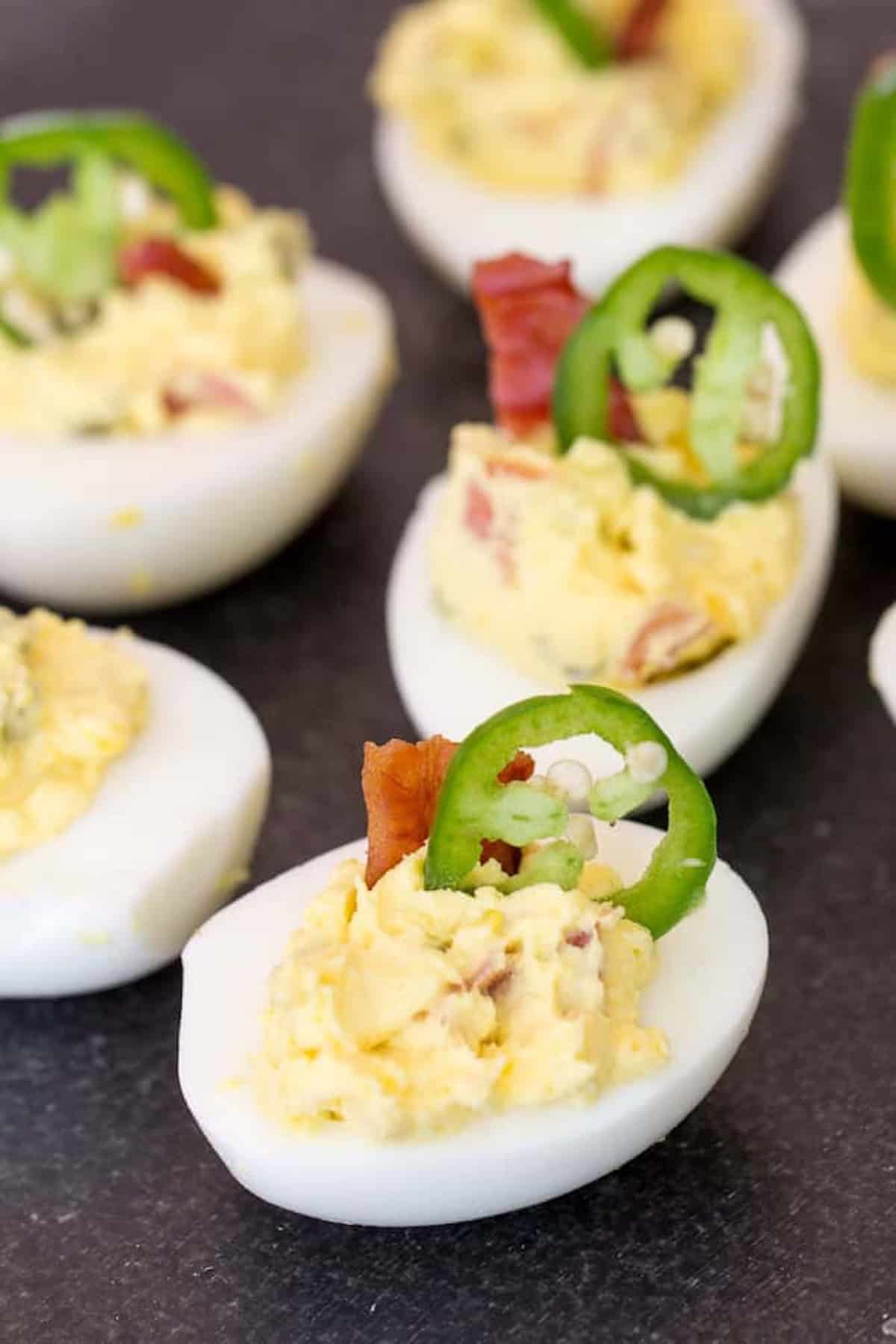 Deviled eggs topped with jalapeño slices and bacon pieces on a dark surface.