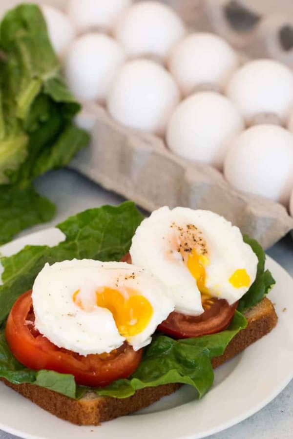 A slice of bread topped with lettuce, tomato, and two poached eggs. Eggs are runny. An egg carton and lettuce are in the background.