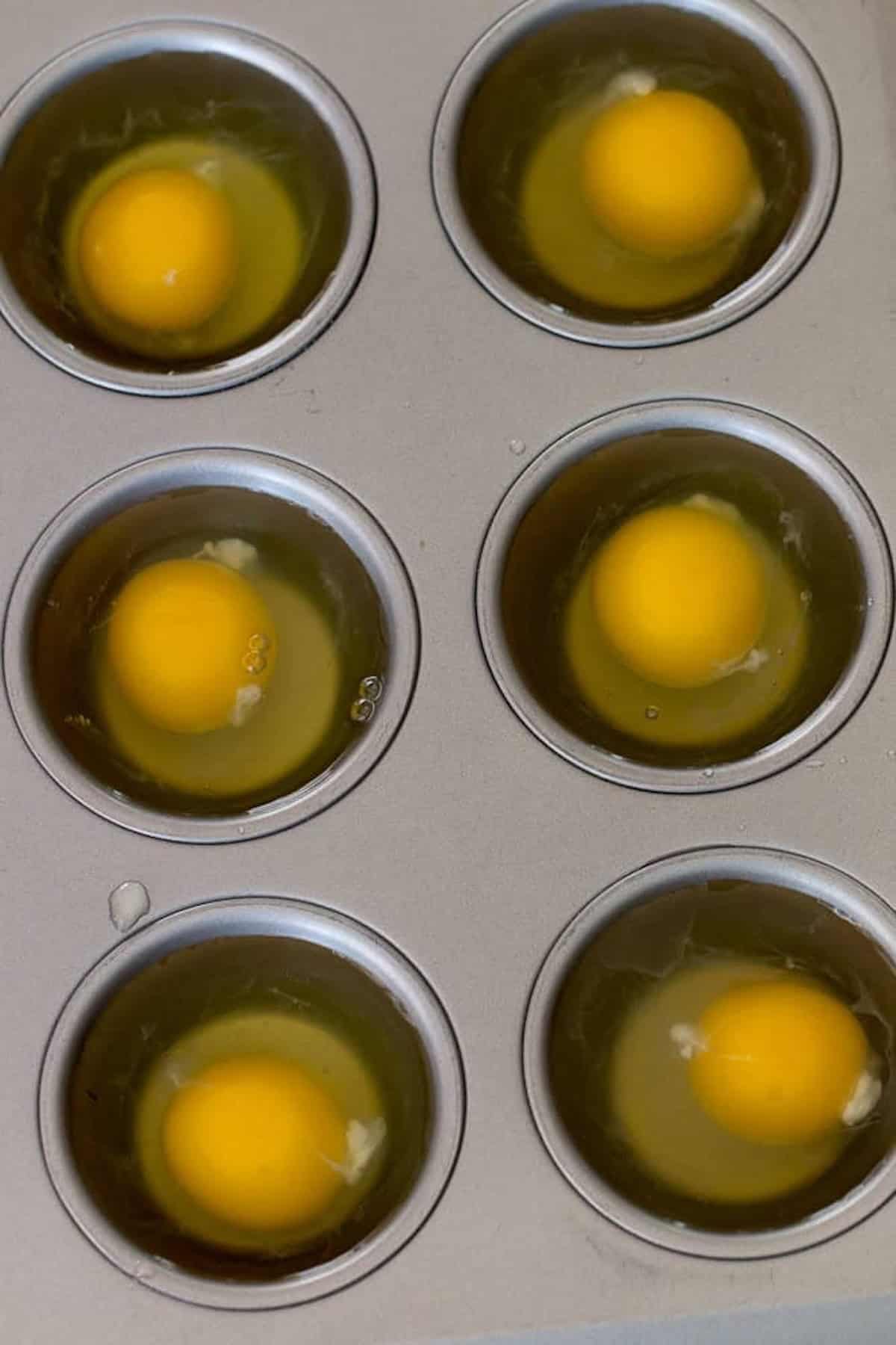 Six raw eggs cracked into a muffin tin, each egg in a separate compartment.