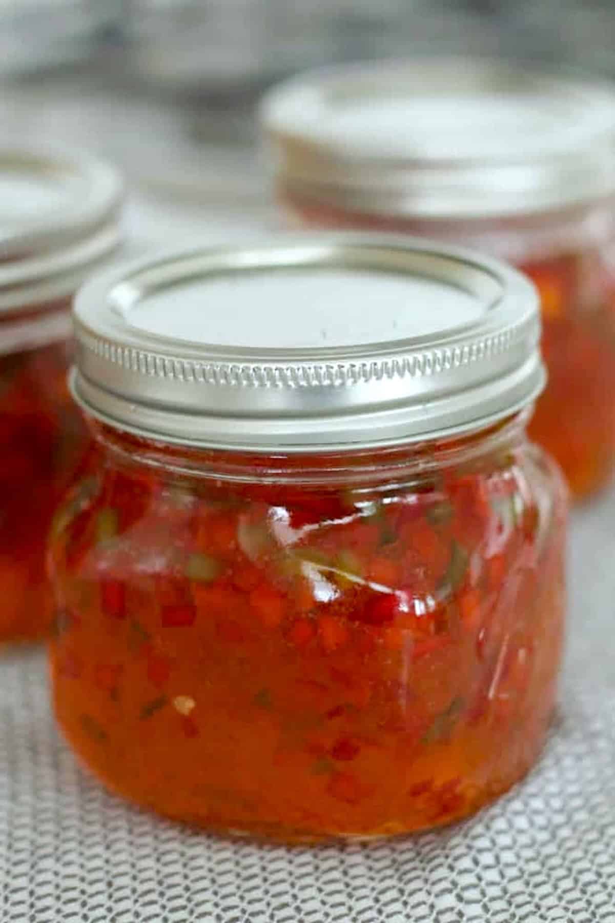 A glass jar filled with red pepper jelly, sealed with a metal lid, sits on a textured surface. Two similar jars are blurred in the background.