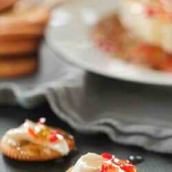 A round cracker topped with a slice of white cheese and red pepper jelly sits on a dark surface, with another similar cracker and a plate in the background.