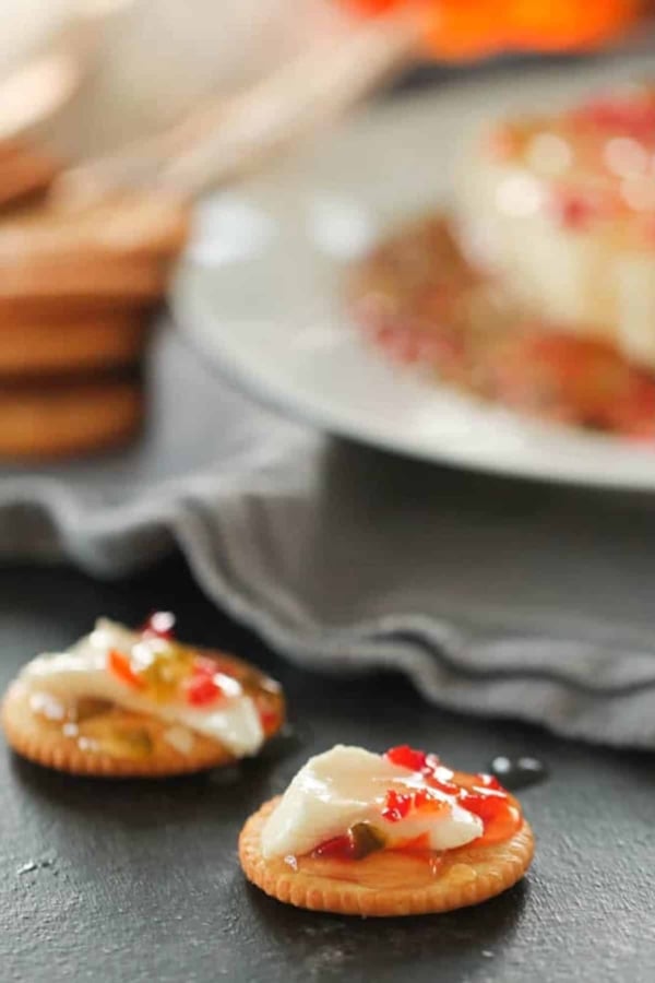 A round cracker topped with a slice of white cheese and red pepper jelly sits on a dark surface, with another similar cracker and a plate in the background.