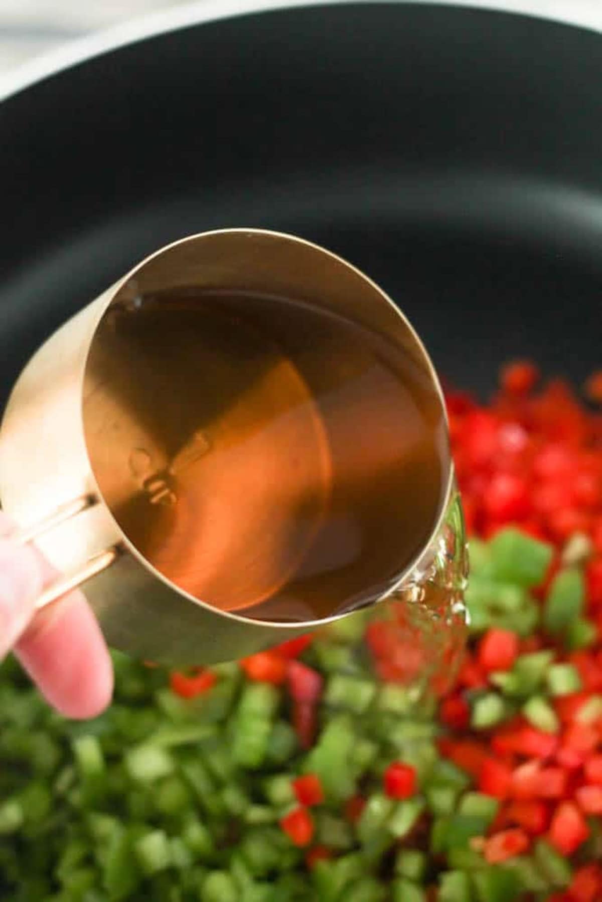 A hand pours liquid from a measuring cup into a pan containing chopped green and red vegetables.
