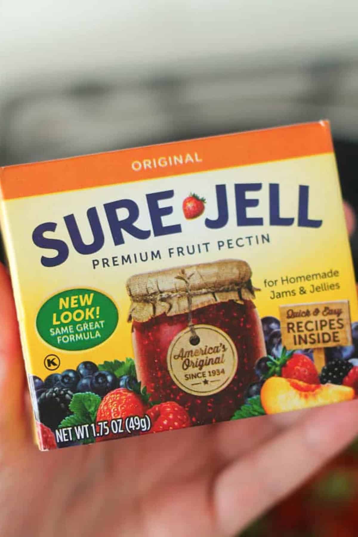A hand holds a box of Sure-Jell Premium Fruit Pectin, labeled for homemade jams and jellies, with images of fruit and a jar of jam on the front.