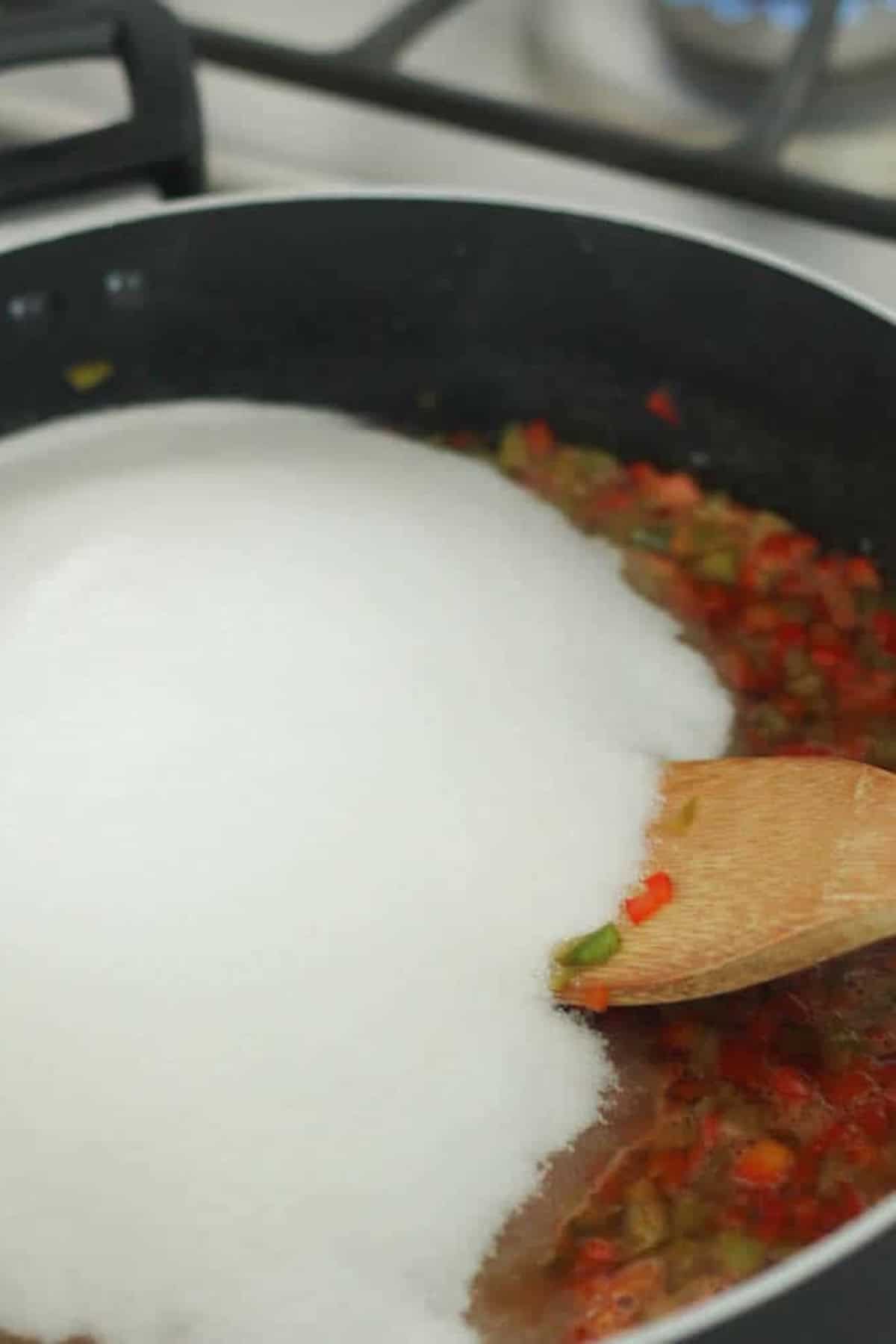 A wooden spoon stirring a large amount of white sugar into a pan of chopped vegetables on a stovetop.