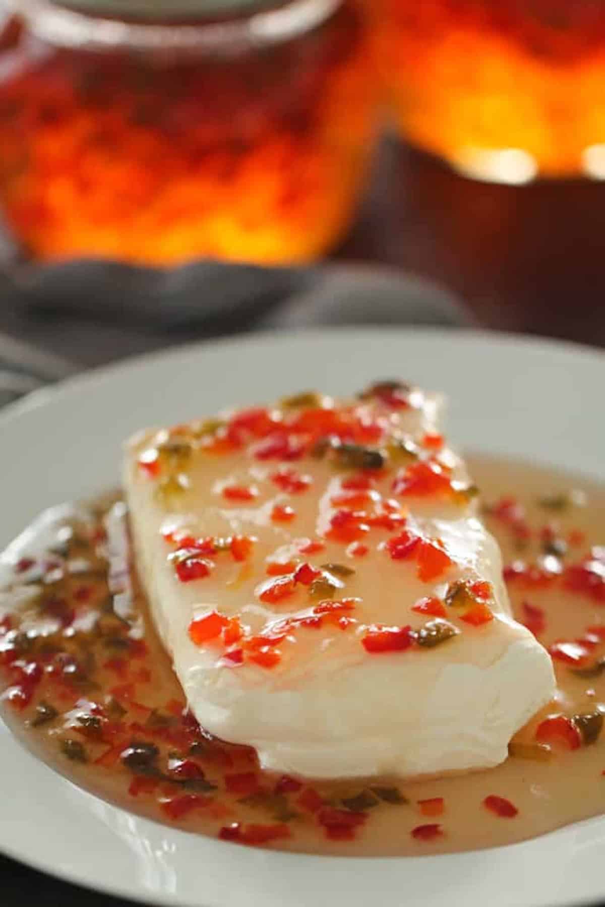 A block of cream cheese covered in a sauce with chopped red and green peppers, served on a white plate.