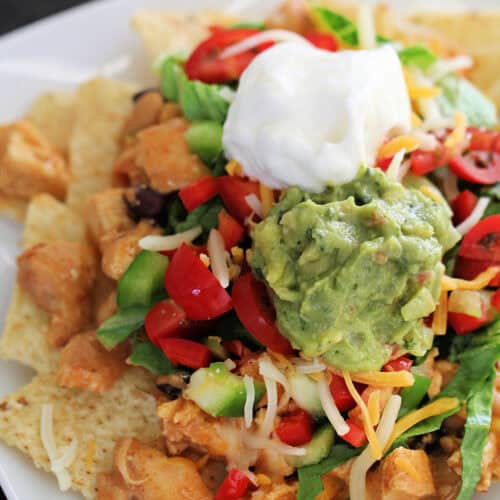 A plate of nachos topped with diced chicken, shredded cheese, lettuce, tomatoes, guacamole, and a dollop of sour cream.