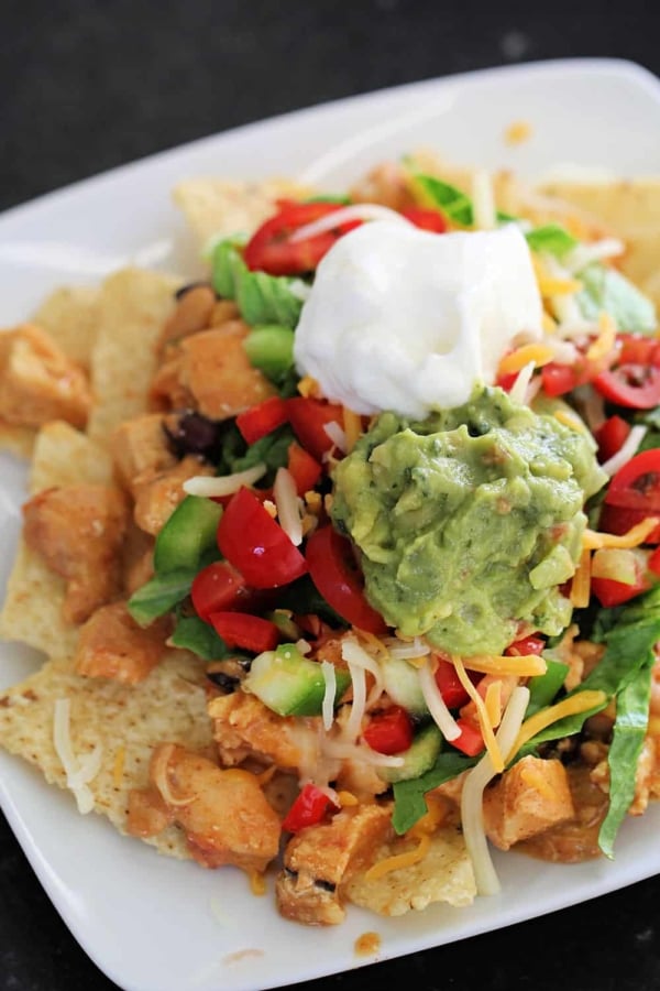 A plate of nachos topped with diced chicken, shredded cheese, lettuce, tomatoes, guacamole, and a dollop of sour cream.