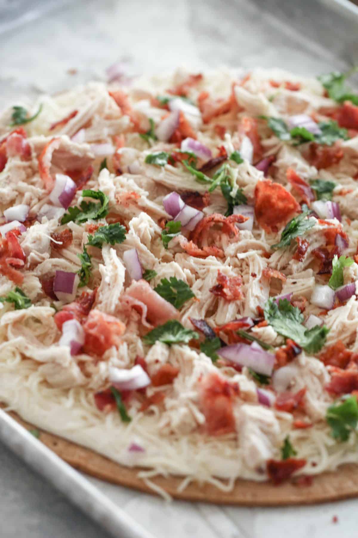 Unbaked pizza topped with shredded chicken, diced tomatoes, chopped red onions, fresh cilantro, and shredded cheese on a baking sheet.