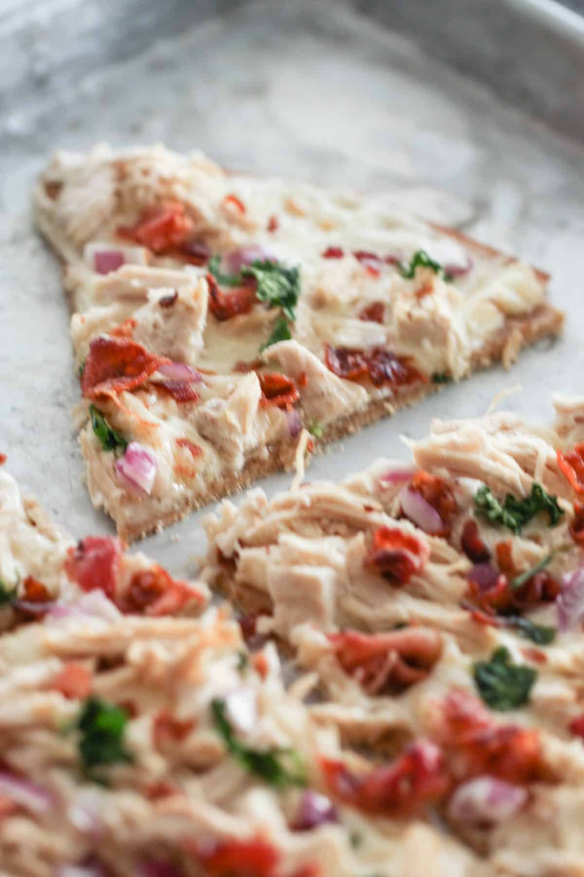 A rectangular pizza slice topped with shredded chicken, red onions, sundried tomatoes, spinach, and melted cheese on a baking tray.