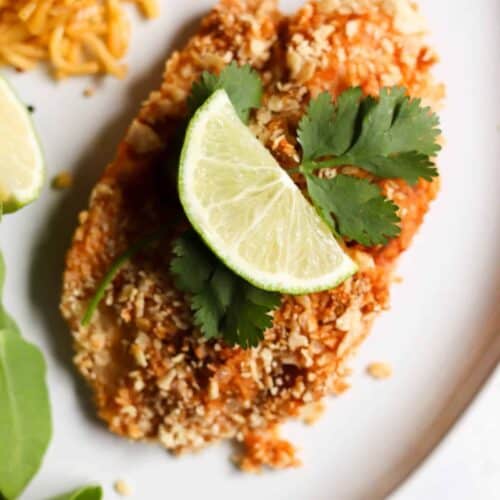 Breaded tilapia garnished with lime slices and cilantro, served on a white plate with a side of rice and greens.
