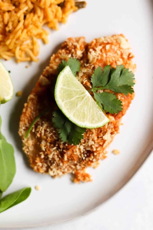 Breaded tilapia garnished with lime slices and cilantro, served on a white plate with a side of rice and greens.