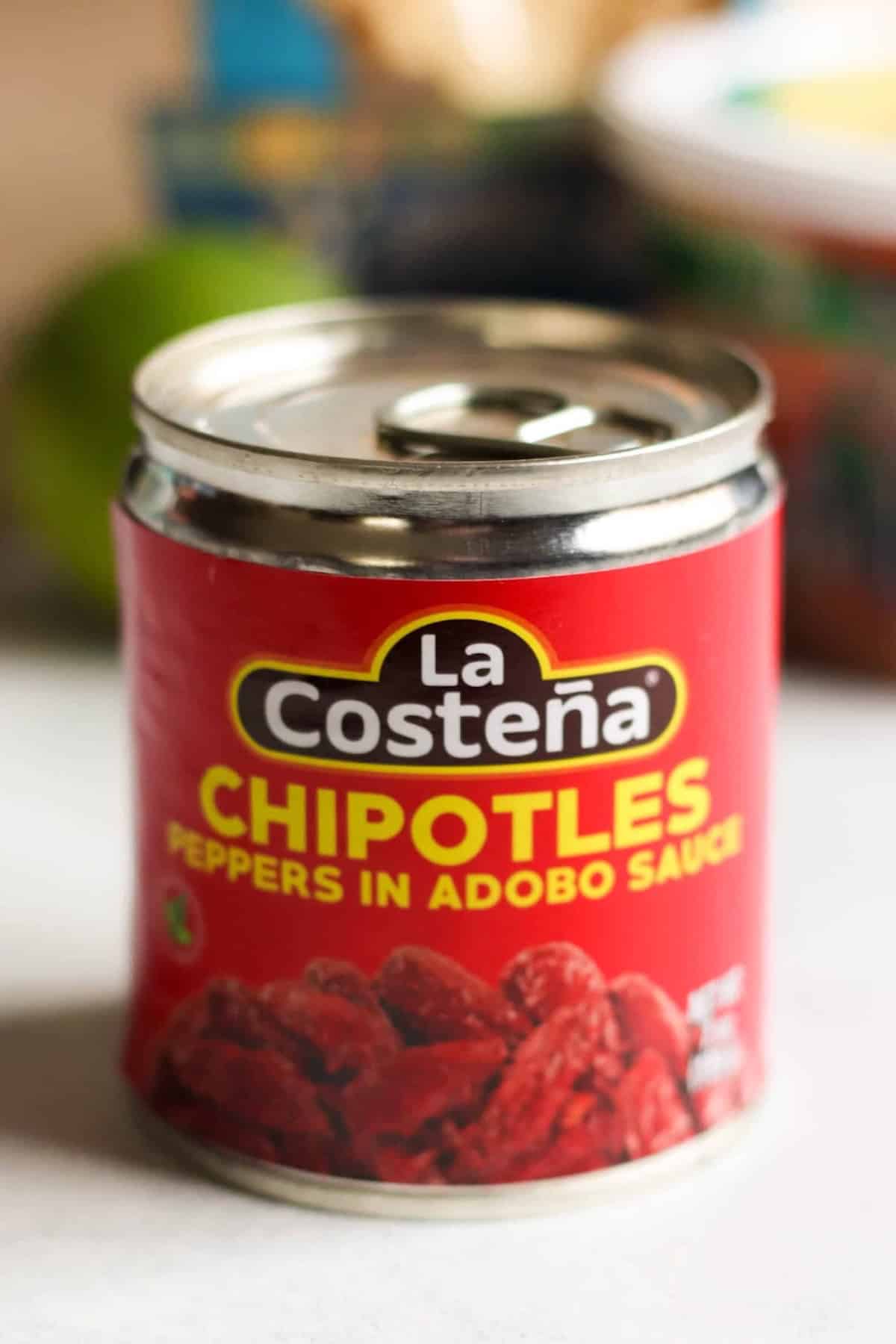 A can of La Costeña chipotles peppers in adobo sauce sits on a countertop, with part of a lime and other blurred objects in the background.