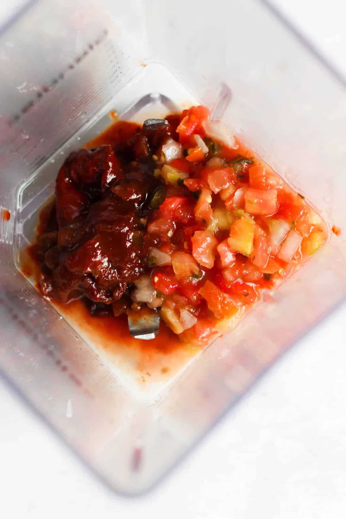 Chopped tomatoes, onions, and chipotle peppers in adobo sauce inside a blender container, viewed from above.