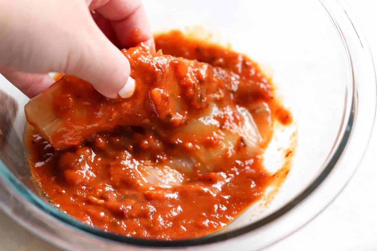 A hand coats a piece of raw tilapia with chunky tomato sauce in a clear glass bowl.