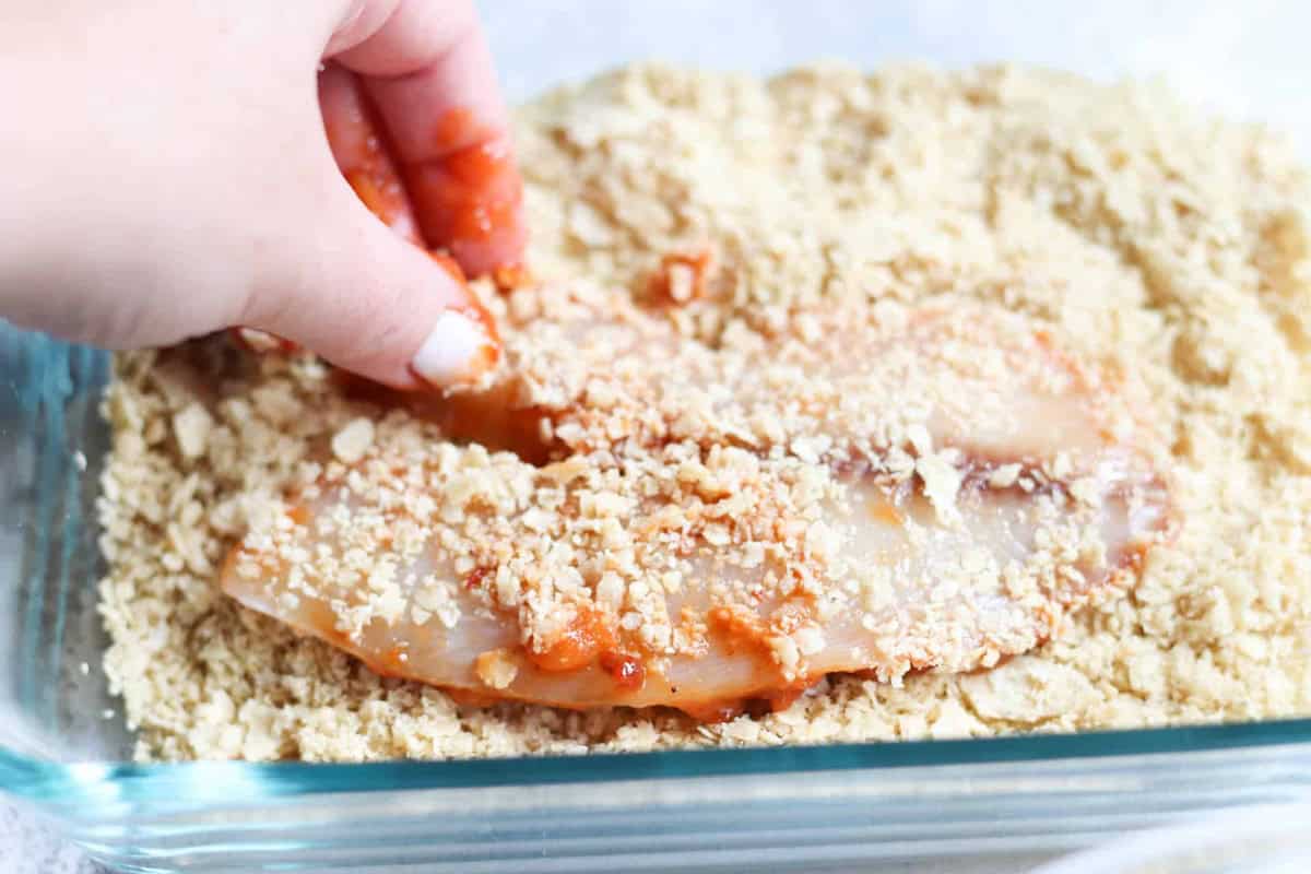 A hand presses a raw, sauce-covered tilapia into a bed of crushed crackers in a glass dish for breading.
