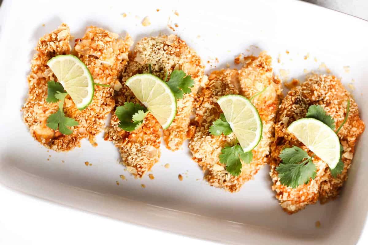 Four pieces of breaded tilapia are garnished with lime wedges and cilantro leaves, arranged in a row on a white rectangular plate.