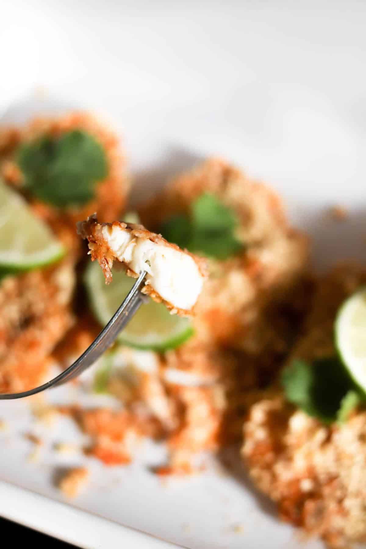 A fork holds a bite of breaded fish fillet, with the rest of the fillets, lime slices, and cilantro in the blurred background on a white plate.