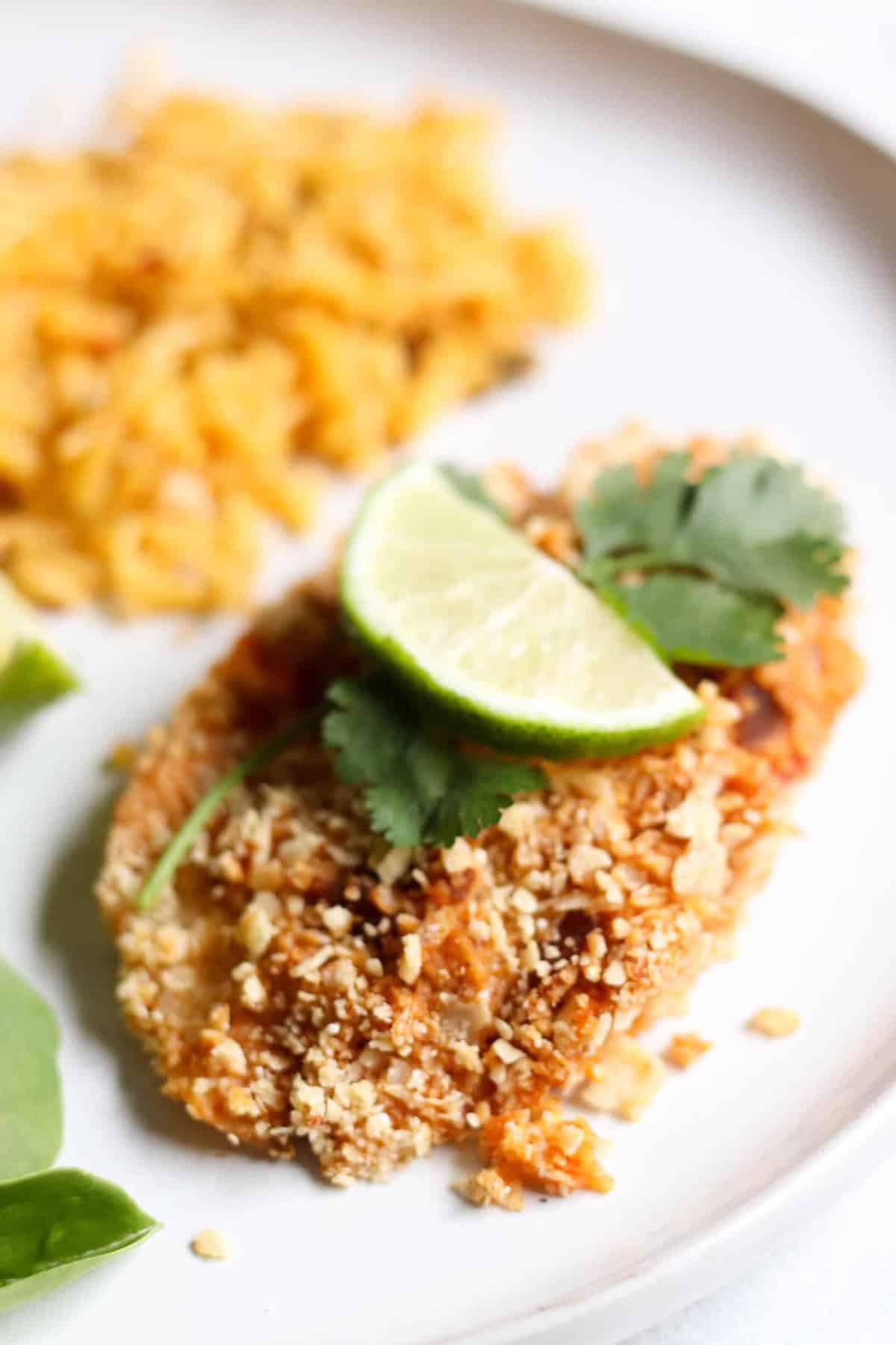 A breaded tilapia garnished with lime slices and cilantro, served on a white plate with a side of seasoned rice.