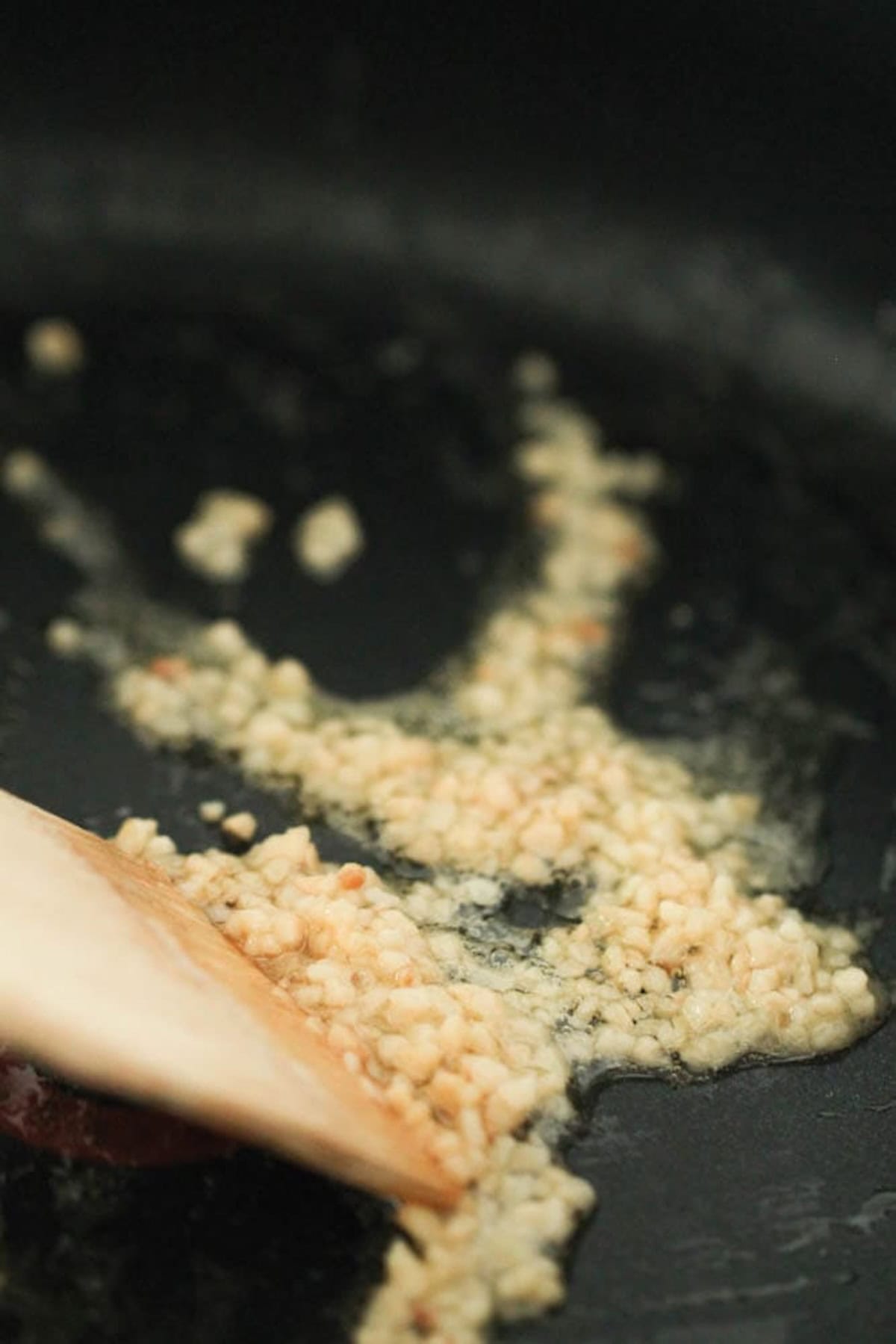 Minced garlic being sautéed in a pan with a wooden spatula.