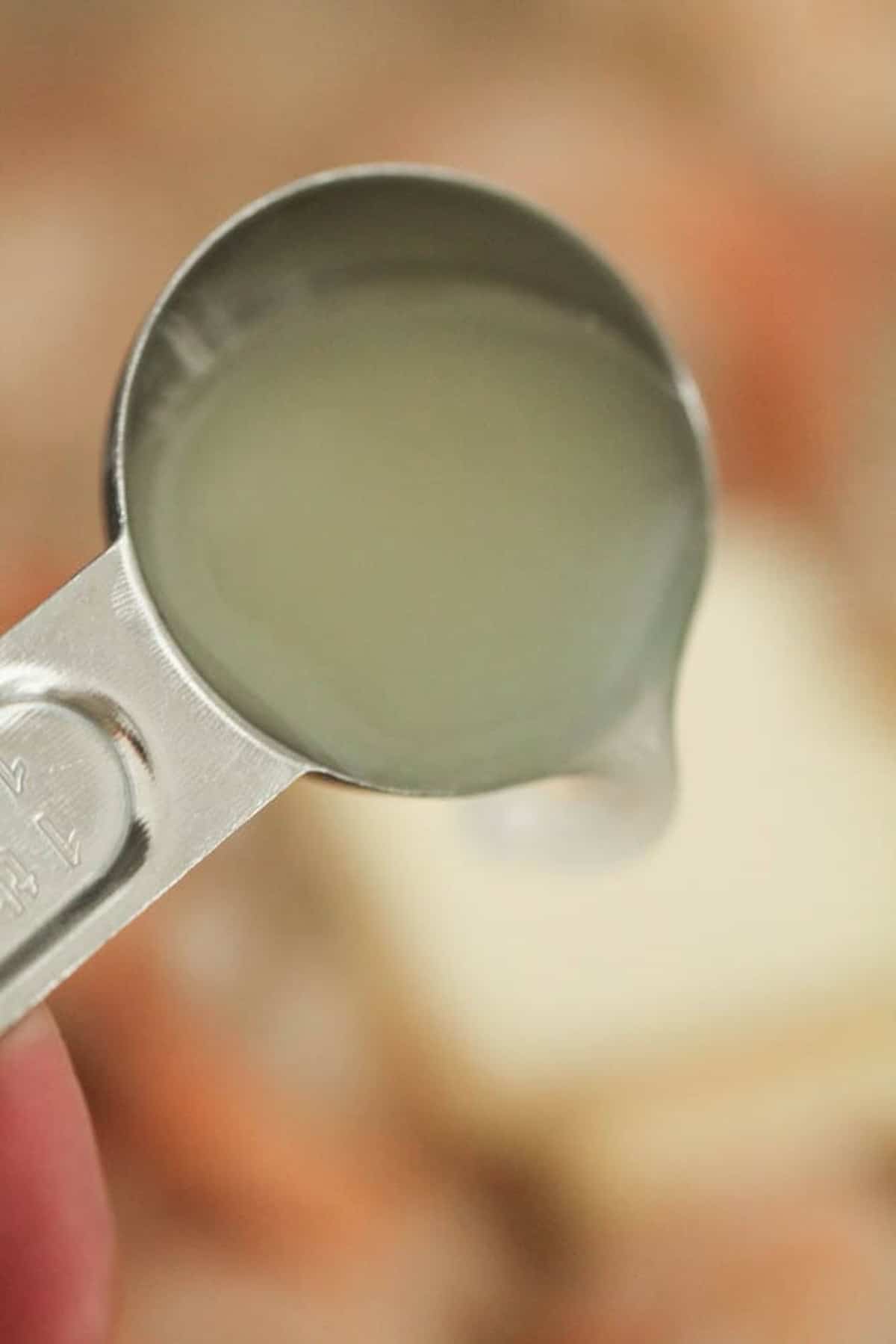 A close-up of a metal measuring spoon filled with a clear liquid, held above a blurred background.